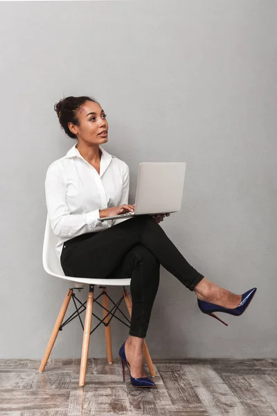 自信的非洲女商人穿着西装坐在椅子上 使用笔记本电脑在灰色 — 图库照片