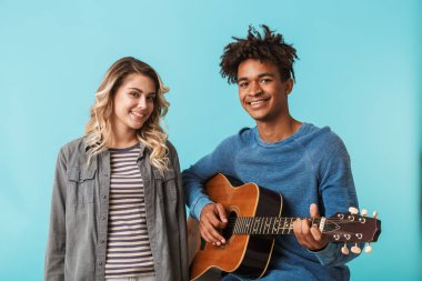 Mutlu genç multiethninc çift birlikte, üzerinde mavi arka plan izole bir gitar çalmak