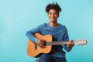 Yakışıklı genç Afrika adam izole mavi üzerinde otururken bir gitar çalmak