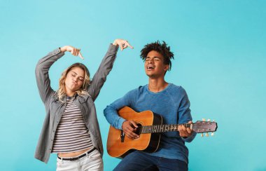 Mutlu genç multiethninc çift birlikte, üzerinde mavi arka plan izole bir gitar çalmak