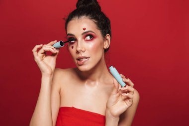 Rimel uygulamak makyaj ayakta poz, kırmızı arka plan üzerinde izole giyen çekici bir genç kadın güzellik portresi