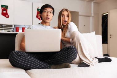 Evde bir kanepede otururken dizüstü bilgisayar kullanan şok genç çift