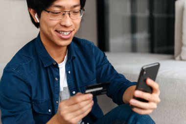 Kulaklık, cep telefonu, kredi kartı tutan kullanarak giymiş evde bir kanepede oturan mutlu Asyalı adam