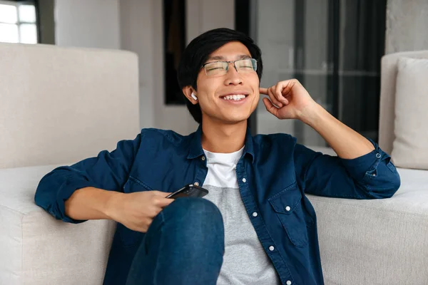 Hombre Asiático Feliz Sentado Sofá Casa Usando Auriculares Sosteniendo Teléfono — Foto de Stock