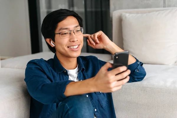 Lycklig Asiatisk Man Sitter Soffa Hemma Bär Hörlurar Hålla Mobiltelefon — Stockfoto