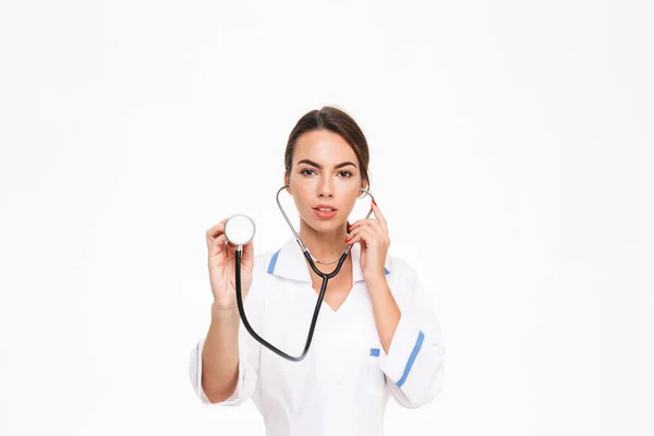 聴診器を使用して制服立っている白い背景に分離された身に着けている美しい若い女性医師 — ストック写真