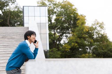 Genç Asyalı adam üstünde hareket eden telefon, açık havada merdiven dururken konuşmaya gülümseyen