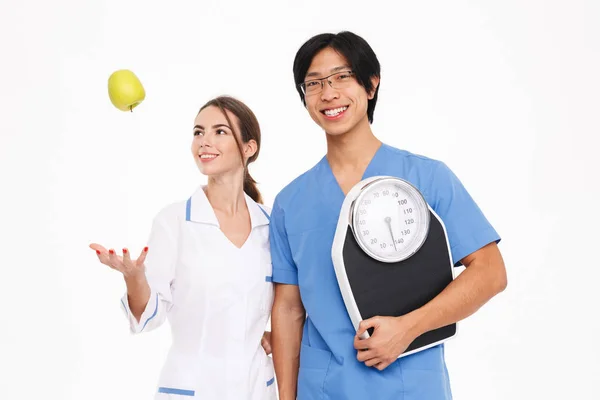 身に着けている白い背景に スケールと青リンゴを保持上分離された制服立っている笑顔の医師カップル — ストック写真