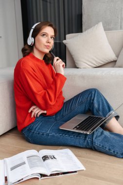 Neşeli genç tesadüfen bir katta evde oturan kulaklıklar dizüstü bilgisayar ile eğitim giyimli kadın