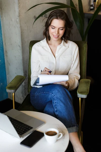ドキュメントを保持のラップトップで屋内で働くカフェに座っている非常に美しい若い女性の画像 — ストック写真