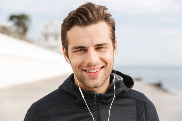 Image of sportive man 30s in black sportswear using earphones and mobile phone while walking along boardwalk at seaside