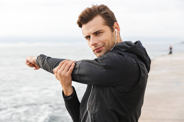 Image of european sportsman 30s in black sportswear and earphones working out at seaside