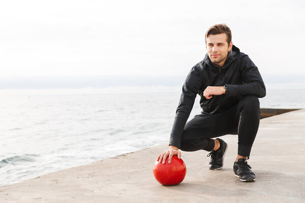 Handsome confident young sportsman working out at the beach, doing exercises with a heavy ball