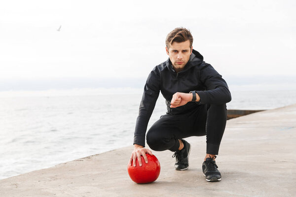 Handsome confident young sportsman working out at the beach, doing exercises with a heavy ball, looking at smartwatch