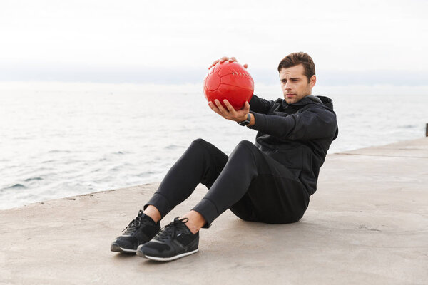 Handsome confident young sportsman working out at the beach, doing exercises with a heavy ball