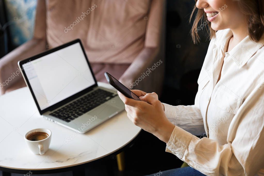 Cropped picture of pretty beautiful young woman sitting in cafe indoors work with laptop using mobile phone.
