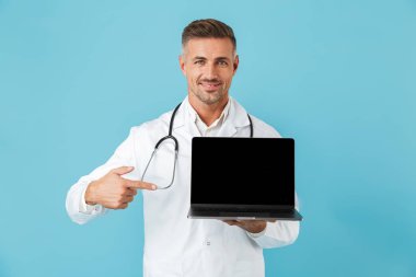 Fotoğraf gülümseyen adam beyaz tıbbi önlük ve mavi arka plan üzerinde izole laptop ayakta tutan stetoskop