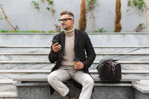 Przystojny Mężczyzna Sobie Kurtkę Trzymając Telefon Komórkowy Siedząc Zewnątrz Picia — Zdjęcie stockowe