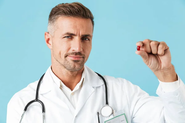 照片中的有吸引力的医生穿着白色外套手拿着一颗药丸站在孤立的蓝色背景 — 图库照片