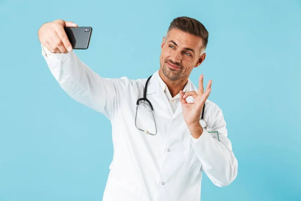 白衣と聴診器の青色の背景に分離立ってのスマート フォン Selfie を取って着て親切な医師の写真 — ストック写真