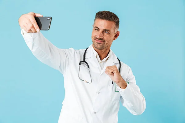 白衣と聴診器の青い背景に分離立っての携帯電話で Selfie を取って着て成熟した医師の写真 — ストック写真