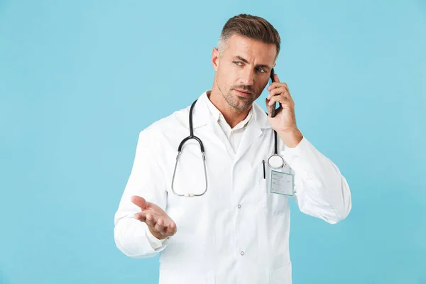 成熟した医師の聴診器の青色の背景に分離立って携帯電話で話していると白衣を着ての写真 — ストック写真