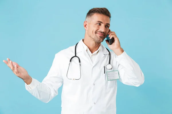 白衣と聴診器の青色の背景に分離立って携帯電話で話しているを着て中年医師の写真 — ストック写真