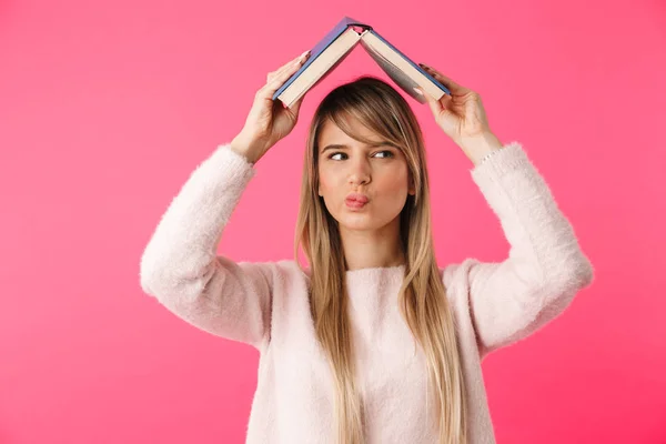 愉快的年轻金发碧眼的女孩站在粉红色的背景孤立 拿着书在她的头上 — 图库照片