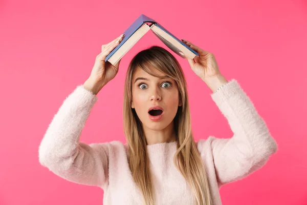 愉快的年轻金发碧眼的女孩站在粉红色的背景孤立 拿着书在她的头上 — 图库照片
