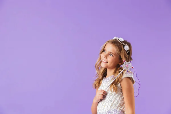 離れて見て 魔法の杖を保持している 紫色の背景に分離されたプリンセスのドレスを着て笑顔のかわいいかわいい女の子の肖像画 — ストック写真