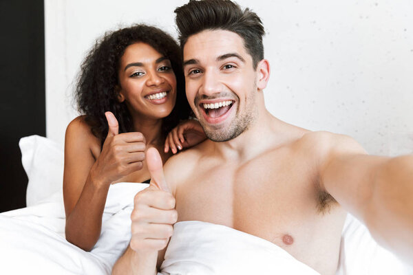 Красивая счастливая молодая многонациональная пара отдыхает в постели под одеялом, делает селфи
