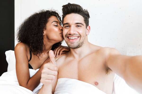 Красивая счастливая молодая многонациональная пара отдыхает в постели под одеялом, делает селфи
