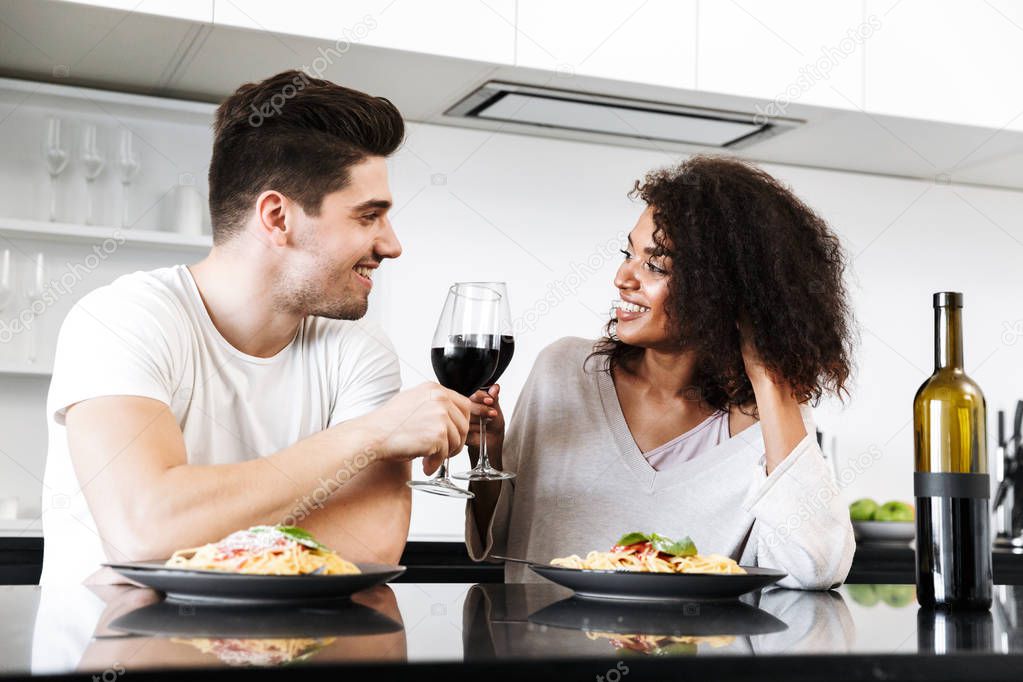 Beautiful young multiethnic couple having a romantic dinner at home, drinking red wine and eating pasta, toasting