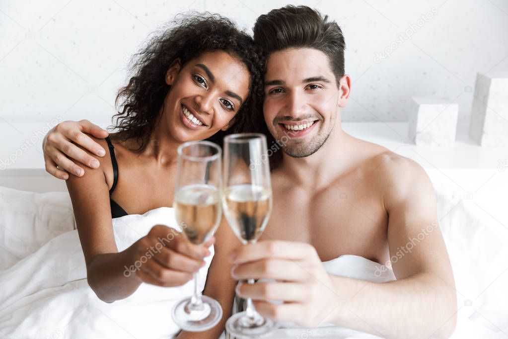 Beautiful happy young multiethnic couple relaxing in bed under blanket, drinking sparkling champagne from glasses