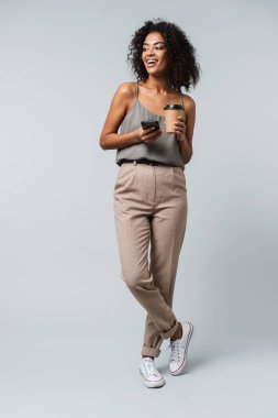 Mutlu bir genç Afrikalı kadın tam uzunlukta raslantı paket servisi olan restoran fincan kahve tutan cep telefonda konuşurken gri arka plan üzerinde izole ayakta giyinmiş