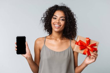 Mutlu genç Afrikalı kadın kayıtsızca giyinmiş gri arka plan üzerinde izole ayakta boş ekran cep telefonu, hediye kutusu açık tutarak