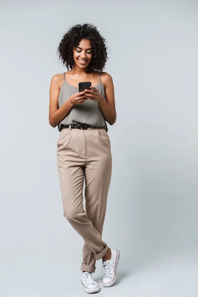 Full length of a happy young african woman casually dressed standing isolated over gray background, holding mobile phone