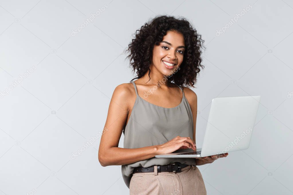 Full length of a happy young african woman casually dressed standing isolated over gray background, working on laptop computer