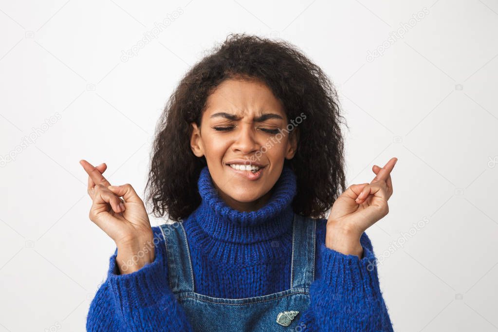 Close up of a pretty worried young african woman wearing sweater standing isolated over white background, holding fingers crossed for good luck