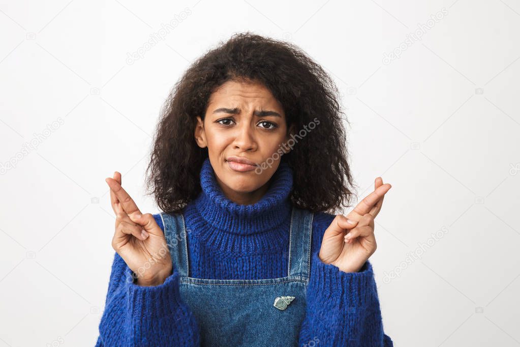 Close up of a pretty worried young african woman wearing sweater standing isolated over white background, holding fingers crossed for good luck