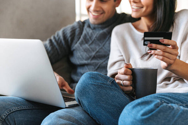 Happy young couple sitting on a couch at home, using laptop computer, showing credit card