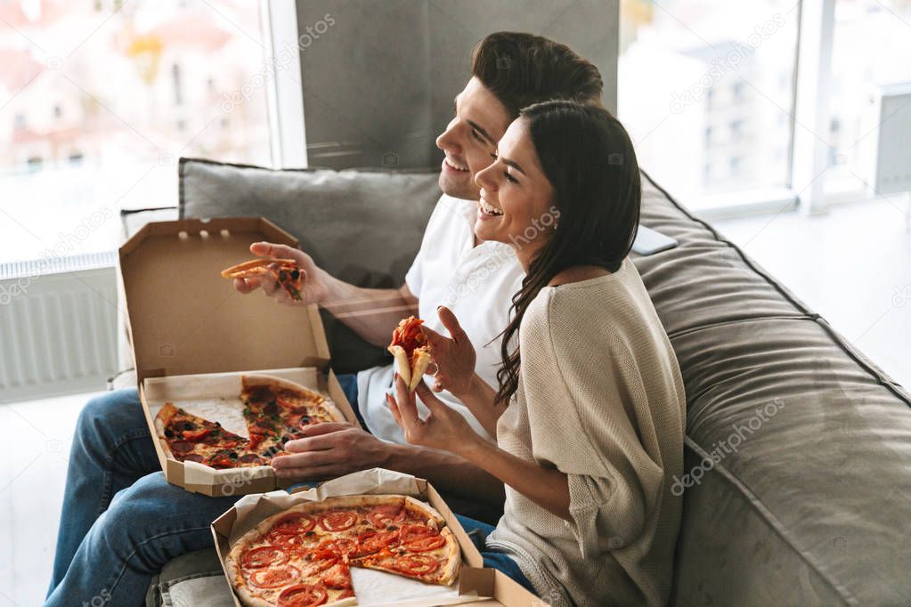 Cheerful young couple sitting on a couch at home, eating pizza, watching TV