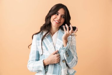 Cep telefonu üzerinde konuşurken bej renkli arka plan üzerinde izole ayakta Ekose ceketli güzel esmer kadın giyinmiş