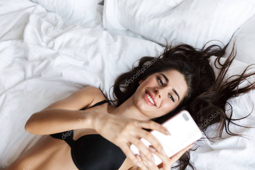 Image of a beautiful brunette woman wearing lingerie using mobile phone lies in bed take a selfie.