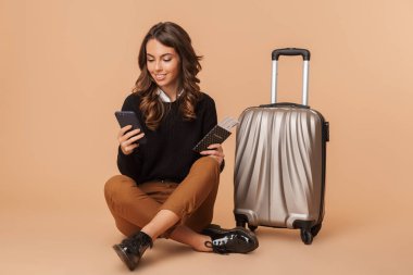 Bej renkli arka plan üzerinde izole katta otururken smartphone ve seyahat bileti tutarak Bagaj ile neşeli kadın