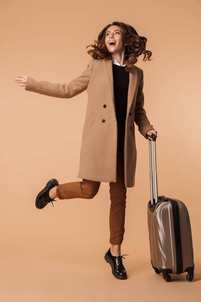 Image of happy woman 20s wearing stylish coat walking with luggage isolated over beige background