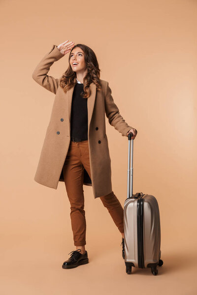 Image of traveler woman 20s wearing stylish coat walking with baggage isolated over beige background