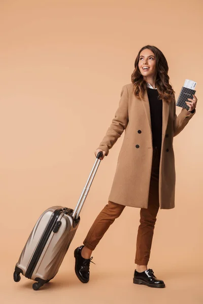 全长一个面带微笑的年轻女子穿着外套走与旅行包查出在米色背景 — 图库照片