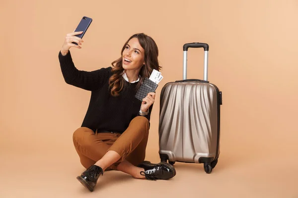带着行李在智能手机上拍摄自拍照片 拿着旅行票的年轻女子 坐在米色背景上隔离的地板上 — 图库照片