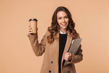 Gülümseyen dizüstü taşıma ve içme kahve bej renkli arka plan üzerinde izole duran bir ceket giyen genç kadın